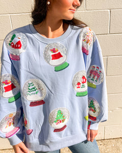 Queen of Sparkles light blue Snow Globe sequin sweatshirt with holiday snow-globe motifs, modeled with jeans outdoors