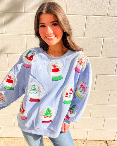 Queen of Sparkles light blue Snow Globe sequin sweatshirt with holiday snow-globe motifs, modeled with jeans outdoors