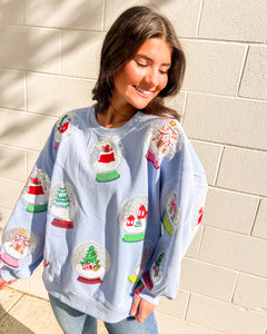 Queen of Sparkles light blue Snow Globe sequin sweatshirt with holiday snow-globe motifs, modeled with jeans outdoors